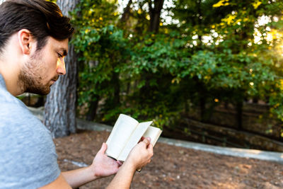 Side view of young man reading book against trees