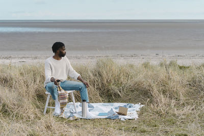 Young man drinking tea while sitting on stool amidst dried plants at beach