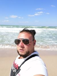 Portrait of young man wearing sunglasses while standing at beach against sky