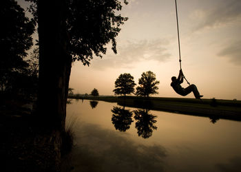 Silhouette man on rope swing over lake during sunset