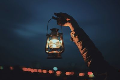 Cropped hand holding illuminated lantern against sky at night