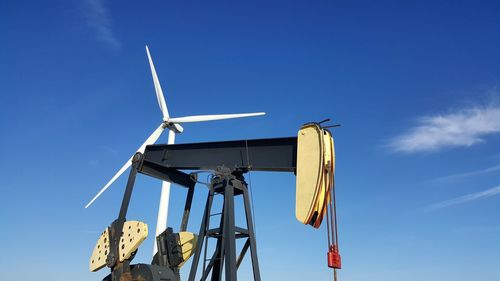 Low angle view of oil drill and wind turbine against blue sky