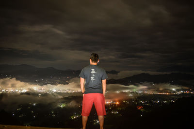 Rear view of man standing on illuminated street against sky at night