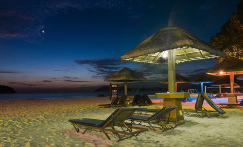 Illuminated parasols with deck chairs at beach against sky at dusk