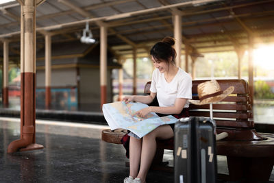 Happy woman with map sitting in bench at railway station