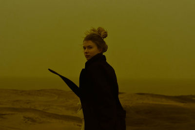 Side view of young woman standing on desert against sky during sunset