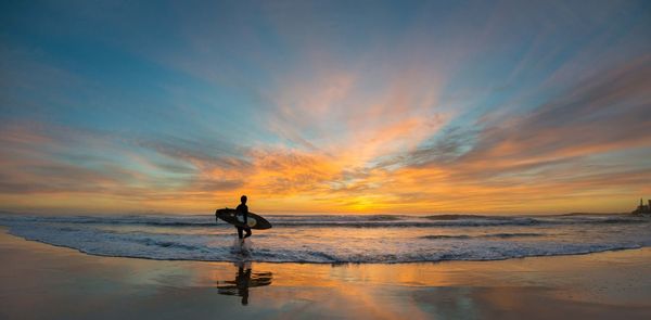 Silhouette man with surfboard walking in sea at beach during sunset