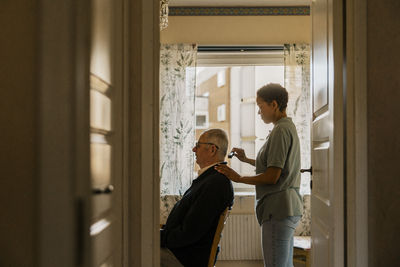 Side view of female nurse combing hair of senior man at home seen through doorway