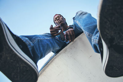 Low angle portrait of man sitting on retaining wall against clear sky