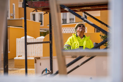 Positive skilled gray haired workman in uniform and protective helmet looking away while standing behind metal construction against building wall in sunny day