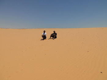 Two men sitting on sand dune in desert against clear sky