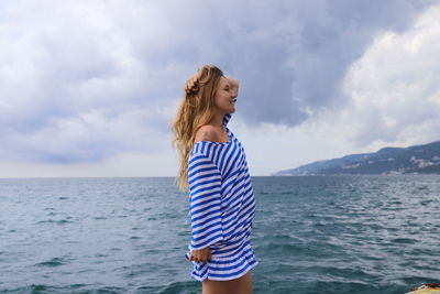 Side view of young woman with hand in hair standing by sea against cloudy sky