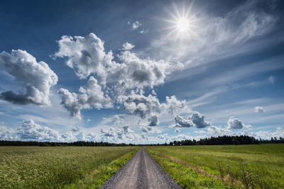 Dirt road amidst green field against cloudy sky during sunny day