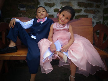 Portrait view of a little boy and a girl wearing formal clothes while sitting on a concrete chair. 