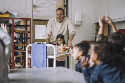 Happy teacher and girl with food trolley by classmates during breakfast in day care center