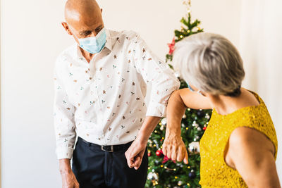 Positive mature couple in smart casual outfits and face masks greeting by bumping elbows while standing near decorated shiny christmas tree