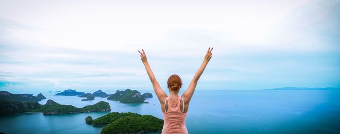 Rear view of woman with arms raised with sea in background against cloudy sky