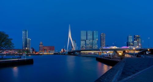 Illuminated modern buildings by river against clear blue sky at night