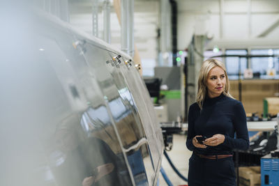 Thoughtful businesswoman with smart phone standing near machine in industry