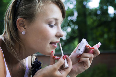 Close-up of young woman applying lip gloss against trees at park