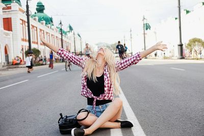 Young woman with arms outstretched sitting on road against sky in city