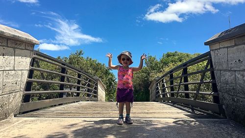 Full length portrait of girl standing on footbridge against sky