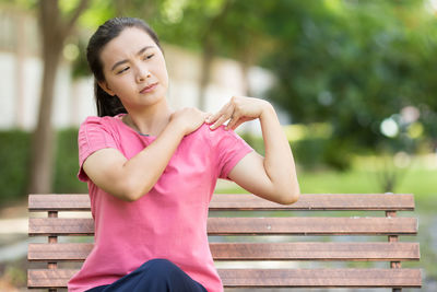 Young woman suffering from shoulder pain while sitting on bench at park