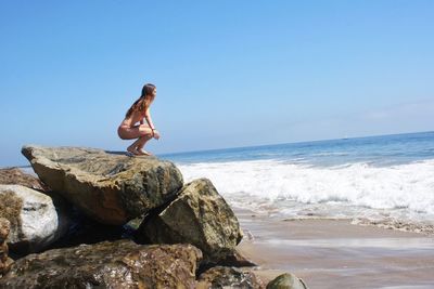 Young woman in bikini crouching on rock formation at beach against sky
