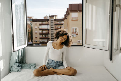 Curly haired woman sitting cross-legged on bed against window at home