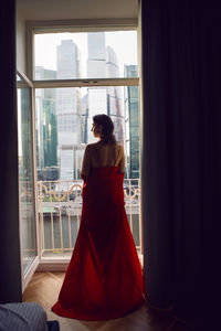 Brunette woman in a red bathrobe stands at a large window in an apartment opposite a skyscraper