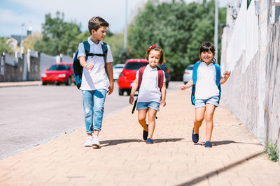 Schoolboy with backpack speaking with female friends while strolling on tiled pavement against stone wall in sunlight