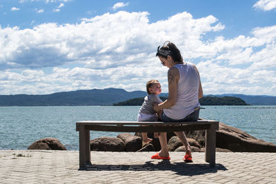 Full length of mother sitting with son on bench at promenade by sea against sky