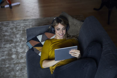 Portrait of smiling woman lying on couch using headphones and tablet