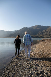 Senior man walking with his granddaughter along the lake shore with mountains.