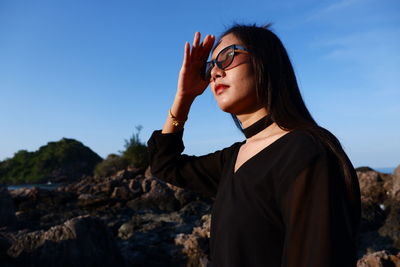 Woman shielding eyes while standing by rock against sky