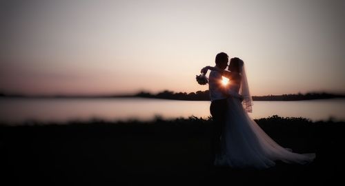 Side view of bride and groom embracing by lake at sunset