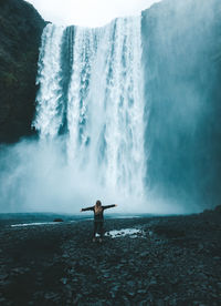 Rear view of woman with arms outstretched standing by waterfall