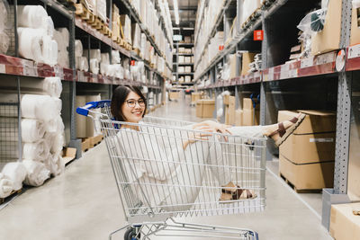 Asian beautiful woman wearing glasses sitting in shopping cart at warehouse furniture shopping store