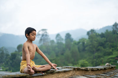 Full length of shirtless man sitting on mountain against sky