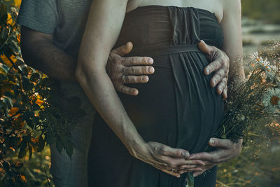 Pregnant woman and her husband hugging her tummy standing outdoors surrounded by nature