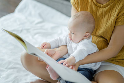 Midsection of mother sitting with baby holding book on bed at home