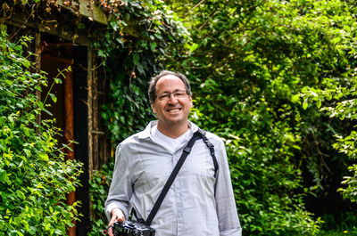 Portrait of smiling man holding camera while standing against trees
