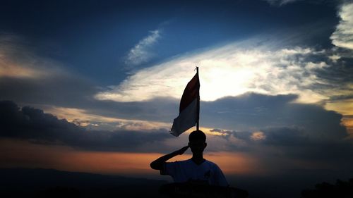 Silhouette man with flag saluting against sky during sunset