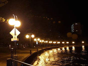 Illuminated street lights by river against sky at night