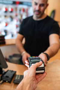 Delighted adult man in black t-shirt smiling and giving payment terminal to crop client while working in bike shop