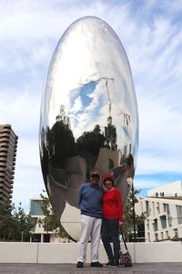 Reflection of man and woman standing on mirror against sky