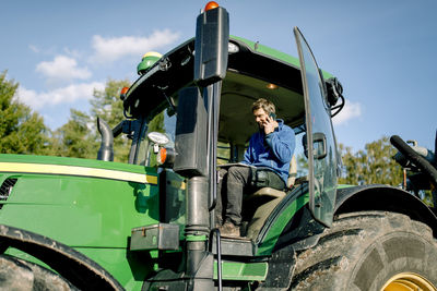 Farmer using tablet pc and talking on smart phone while sitting in tractor at farm