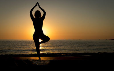 Silhouette woman meditating while standing on shore at beach against sky during sunset