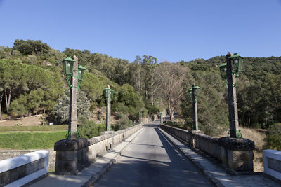 Empty road along plants and trees against clear blue sky