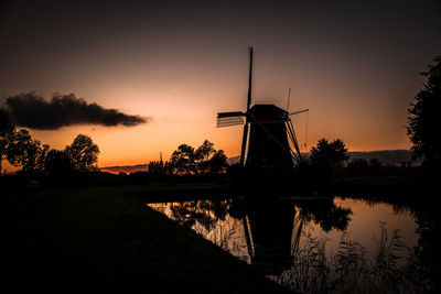 Silhouette traditional windmill by lake against sky during sunset
